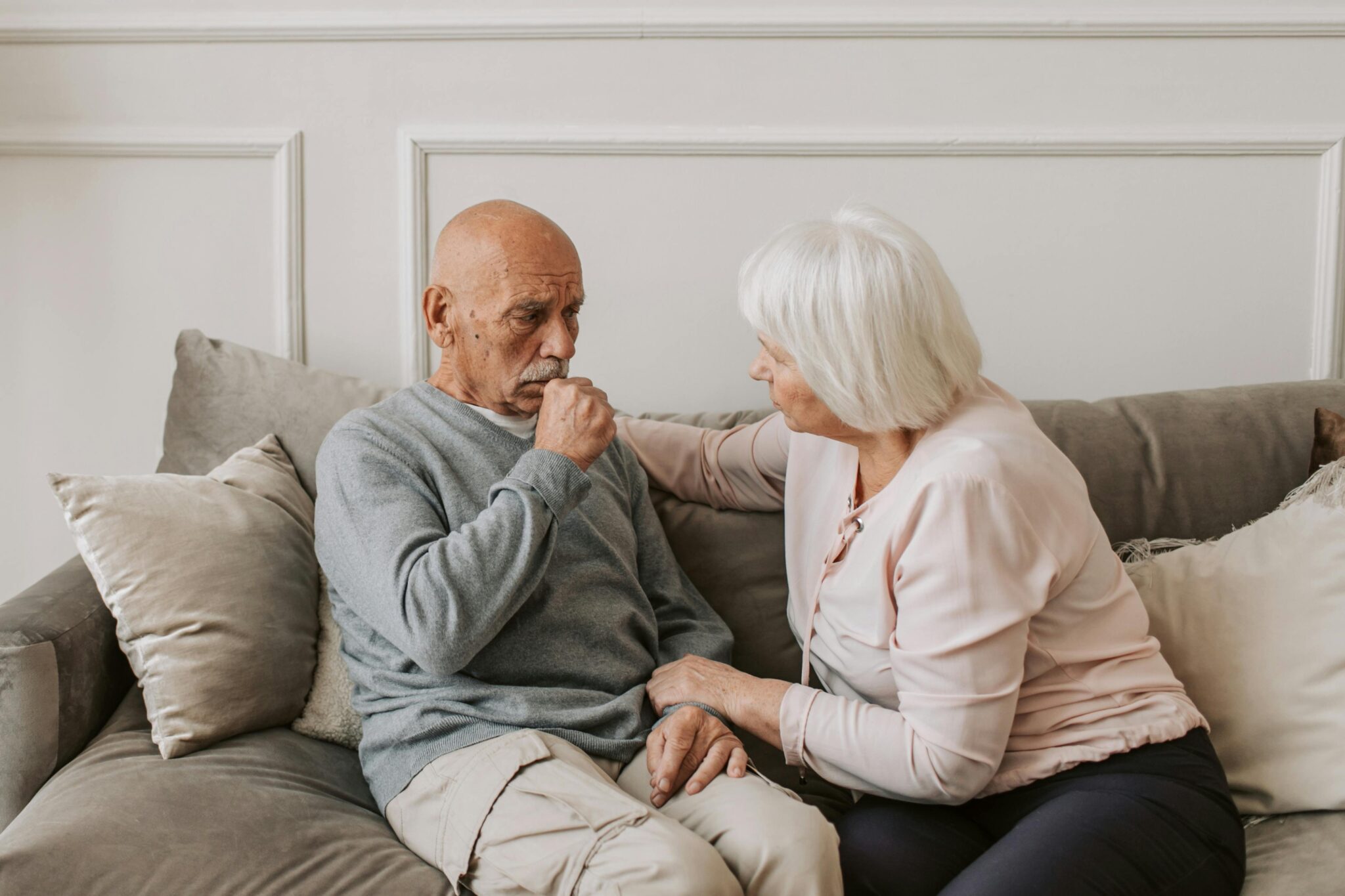 Older man and woman sitting on couch with woman's hand on the shoulder of man while he covers his mouth with a fist as if to be short of breath or coughing.