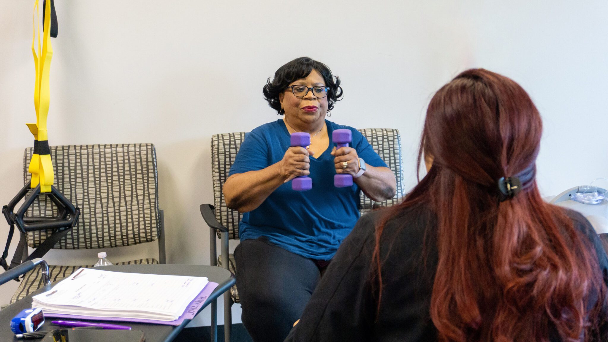 Woman sitting down holding weights with bent elbows and looking across at a female pulmonary rehab technician.